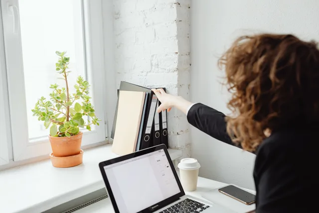 A woman reaches for folders in a light-filled modern office with a laptop. Große Dateien sicher an Mitwirkende übergeben