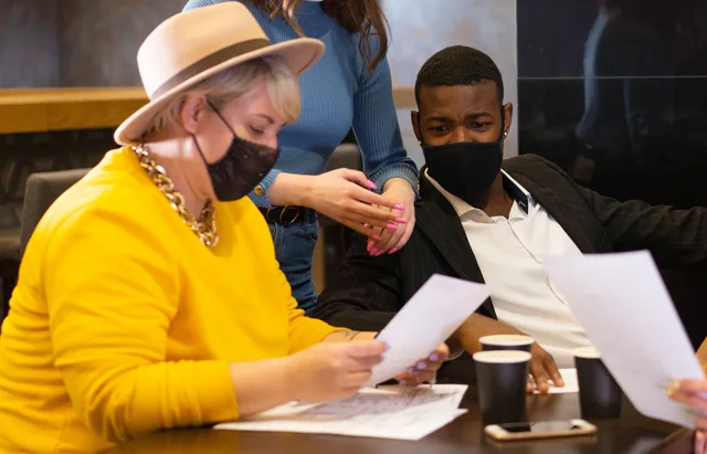 Diverse group discussing documents with disposable cups and face masks indoors. Teamarbeit in Creator-Projekten
