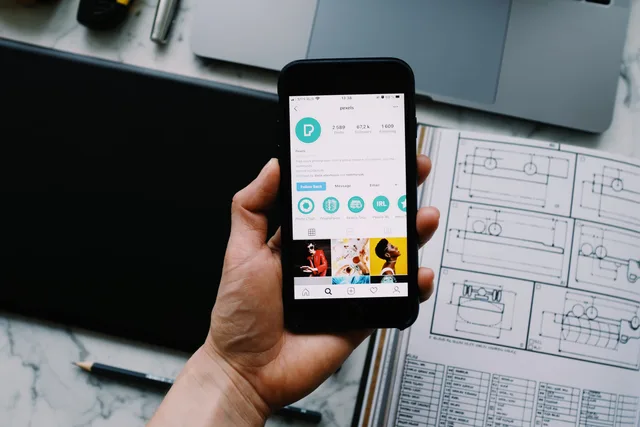 Hand holding smartphone displaying social media profile with tech gadgets on desk. Publishing-Zugriffe auf Social-Konten begrenzen